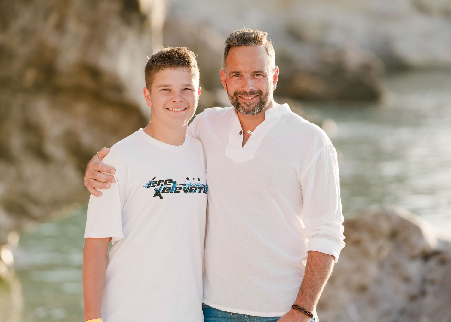 Two people pose together on a sunny beach with coastal rock formations and blue water in the background. Fotograf Rhodos