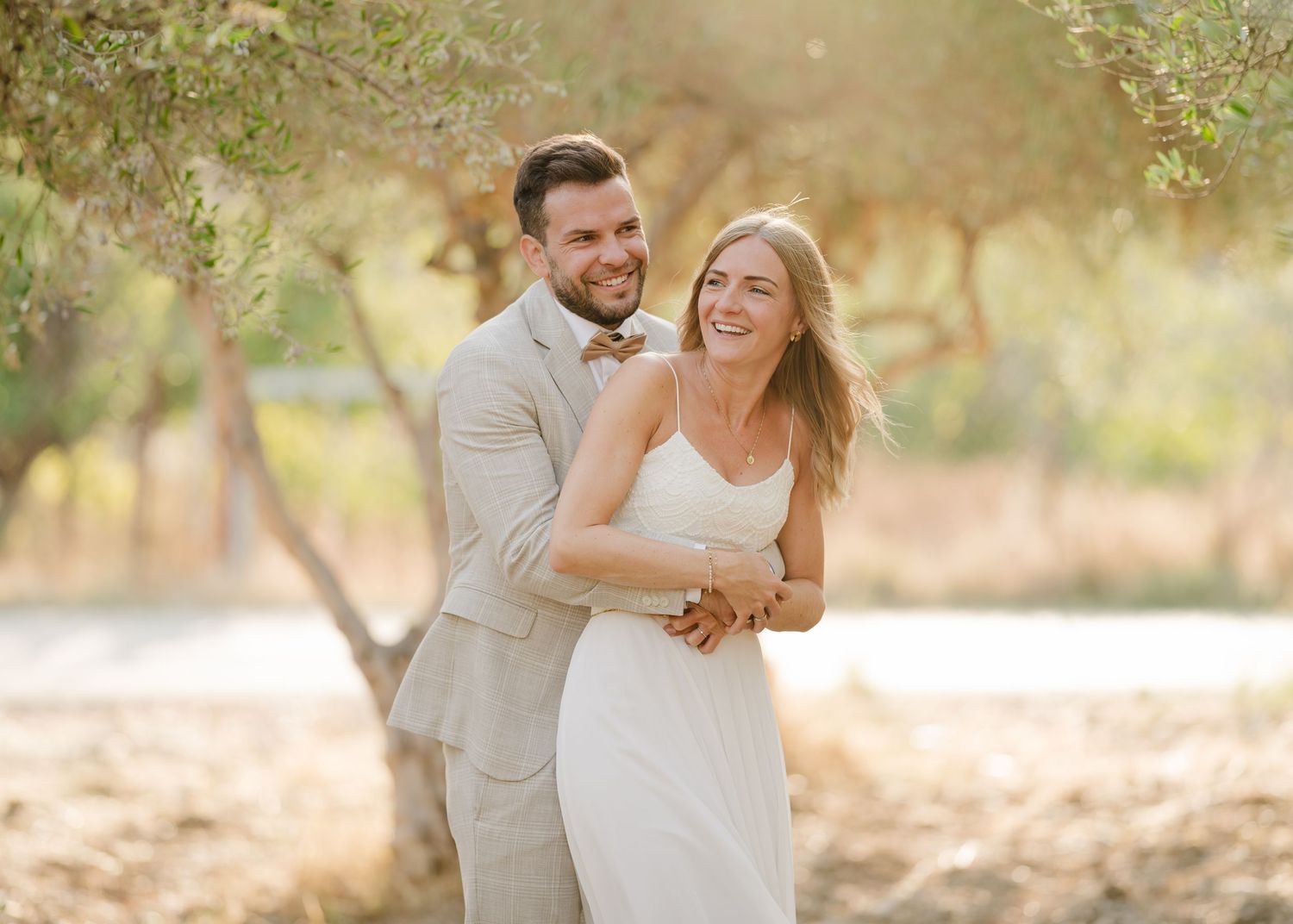 Das Paar umarmt sich freudig im Freien mit sanften, sonnenbeschienenen Bäumen im Hintergrund. Hochzeiten Griechenland - Hochzeitsfotograf Rh