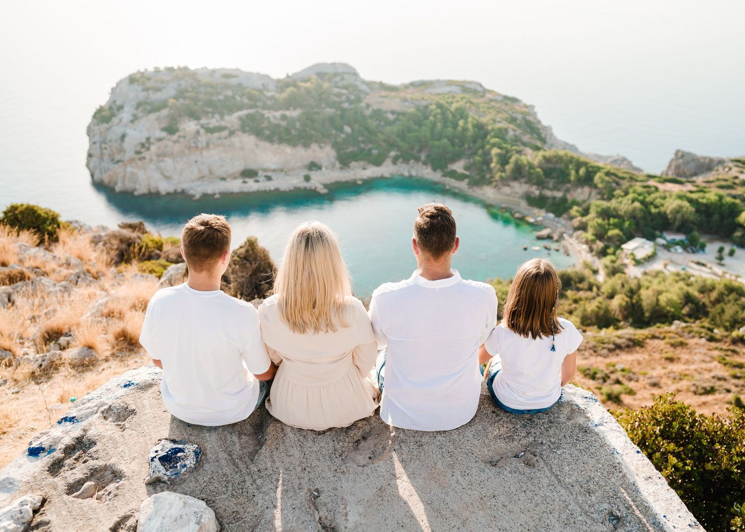 Family sitting at cliff edge overlooking spectacular coastal view with turquoise water bay below. Fotograf Rhodos