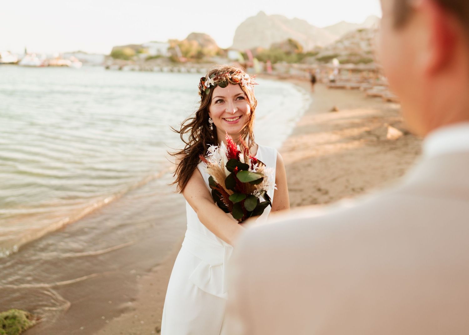 Braut mit Blumenstrauß lächelt am Strand mit warmer Sonnenuntergangsbeleuchtung.