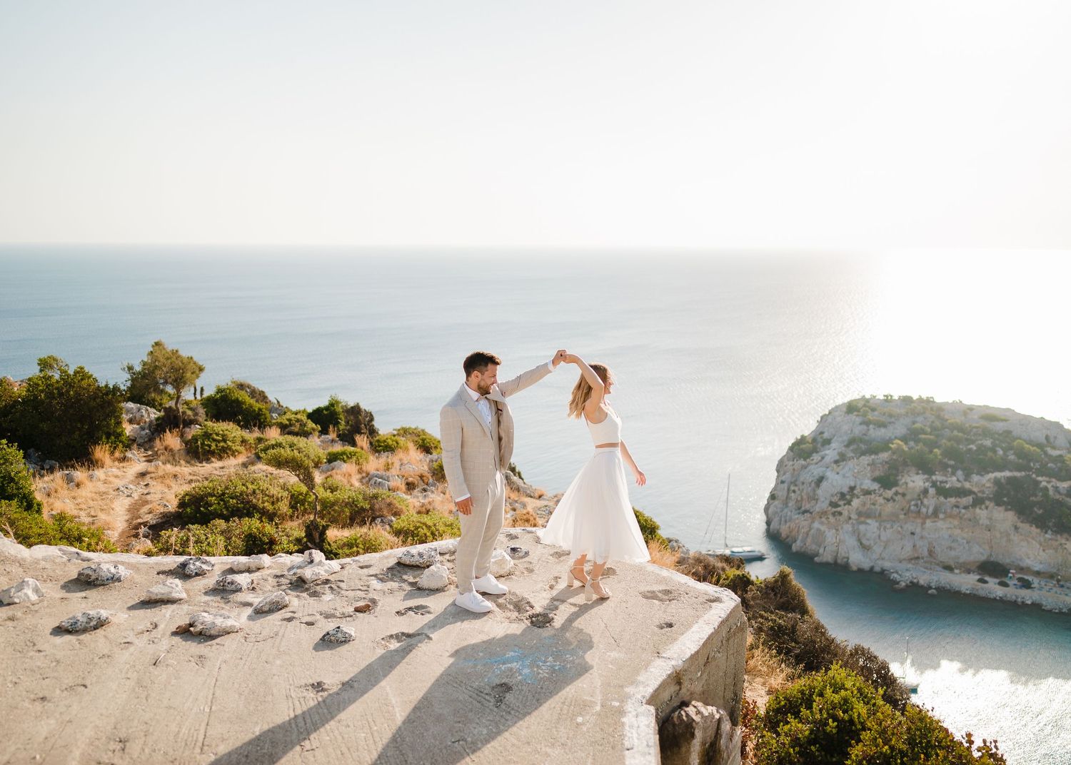 Das Paar tanzt auf einem malerischen Aussichtspunkt mit dramatischem Küstenblick und klarem blauem Wasser darunter. Strandhochzeit Rhodos -