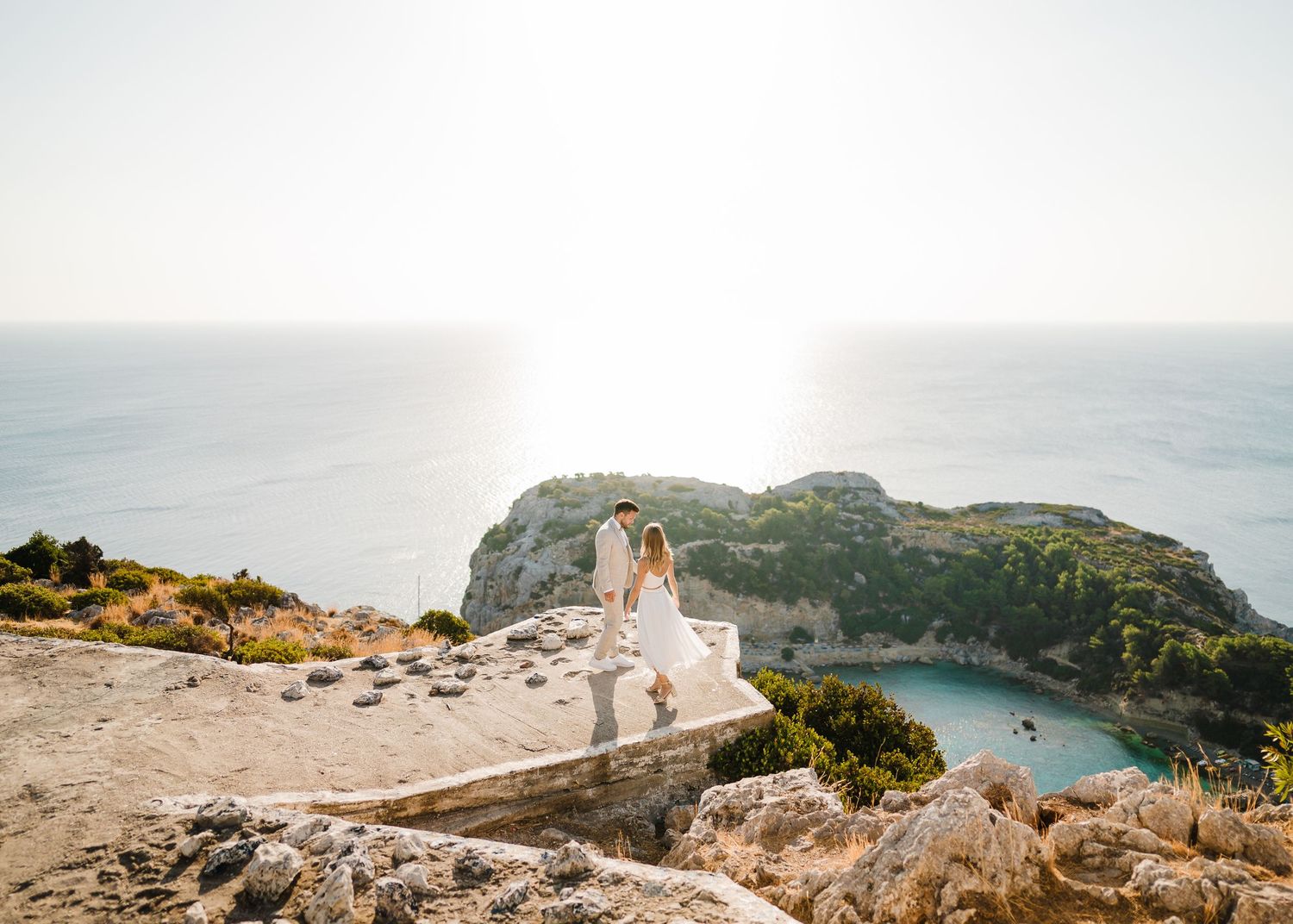 Das Hochzeitspaar steht auf einer dramatischen Klippe mit Blick auf die Mittelmeerbucht und das kristallklare Wasser. Strandhochzeit Rhodos