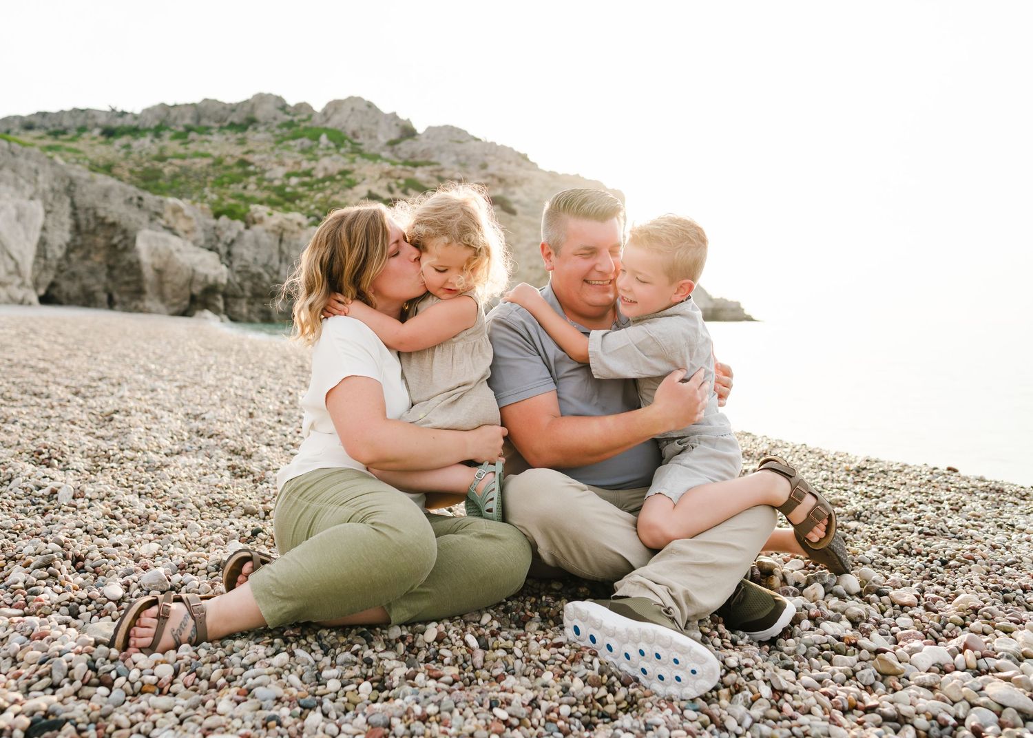 Family embraces and smiles while sitting together on pebble beach near cliffs. Fotograf Rhodos