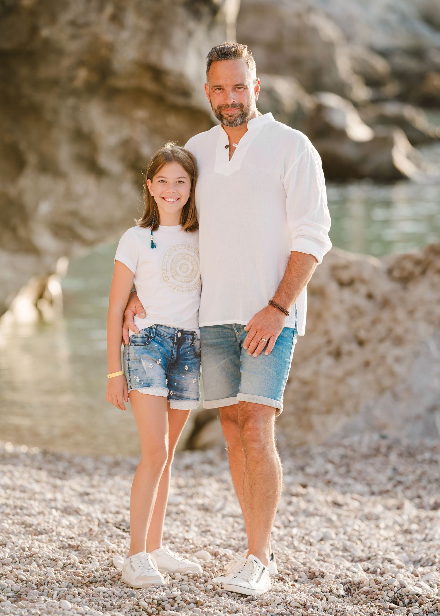Two people stand together on a pebbly beach with dramatic coastal cliffs and sparkling water behind them. Fotograf Rhodos