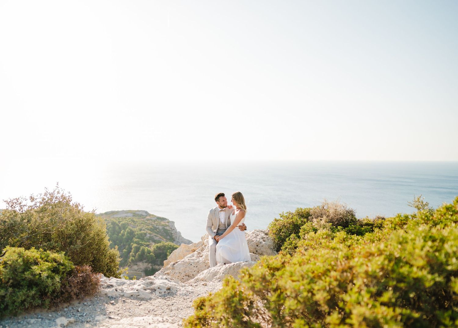 Paar in Hochzeitskleidung steht an einem malerischen Küstenweg, umgeben von mediterraner Vegetation. Fotografin Rhodos - Hochzeitsfotograf R