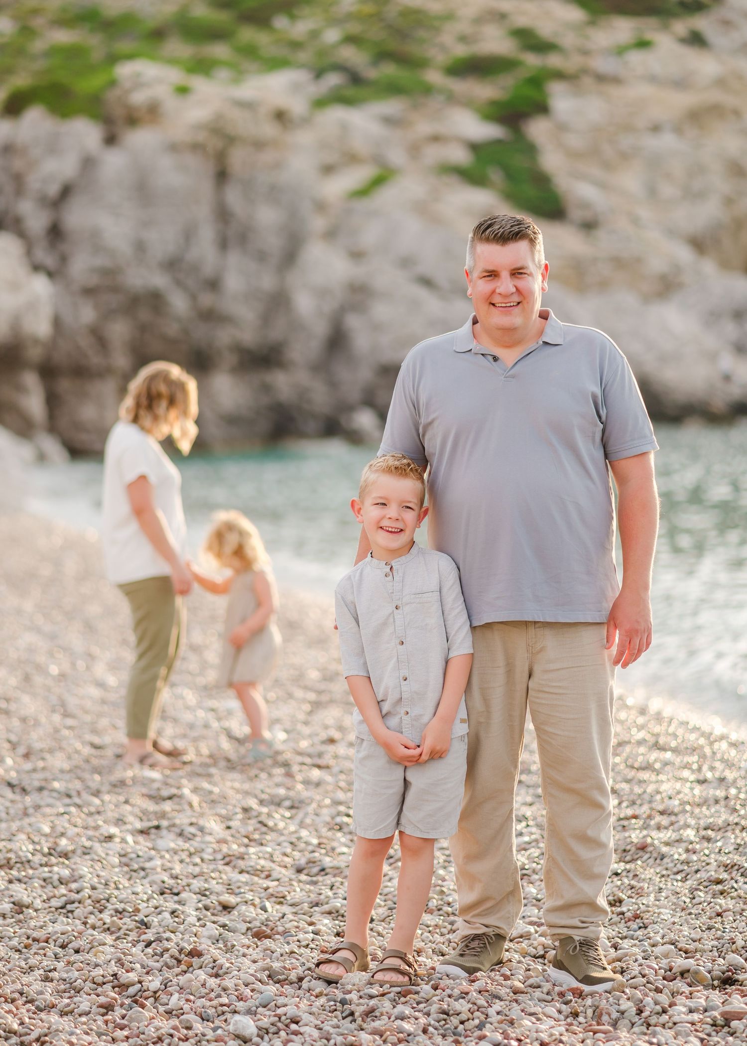 Family members posing on pebbly beach with coastal rocks and turquoise water in background. Fotograf Rhodos
