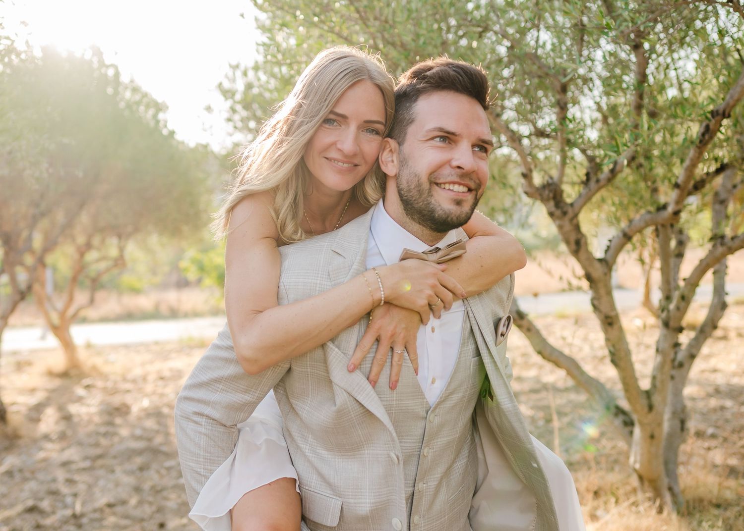 Freudiges Paar in leichter Hochzeitskleidung mit verspieltem Huckepack-Moment zwischen Olivenbäumen bei Sonnenuntergang. Hochzeiten Griechen