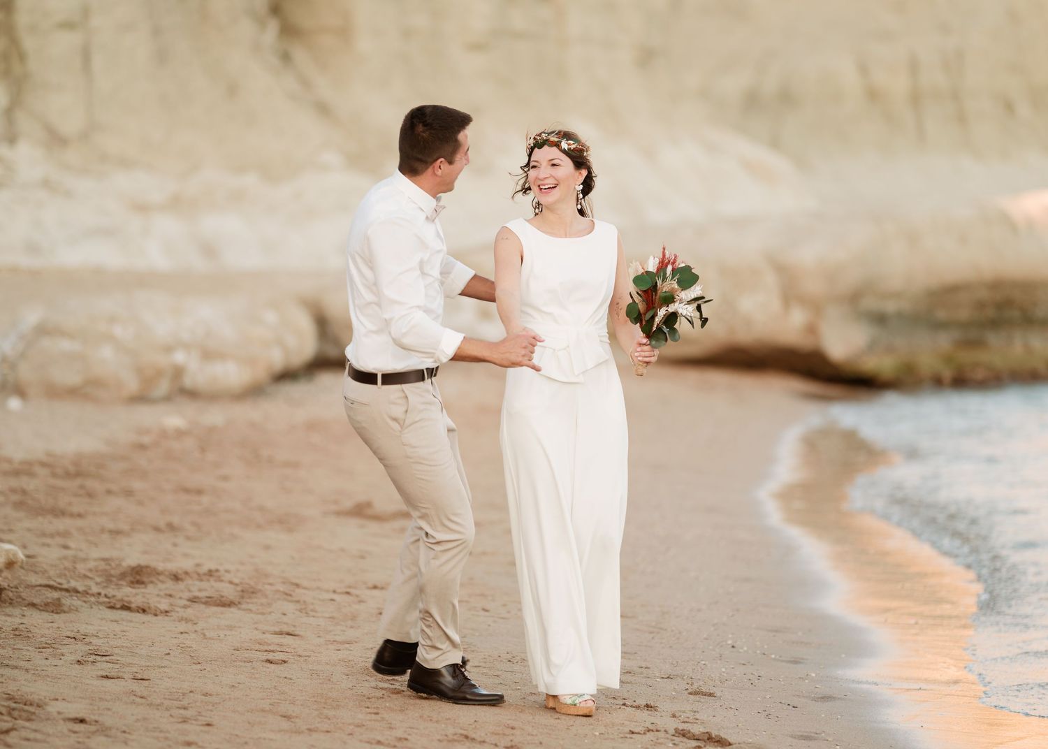 Hochzeitspaar, das einen verspielten Moment am Strand mit Klippenhintergrund teilt. Hochzeiten Griechenland - Hochzeitsfotograf Rhodos Tina