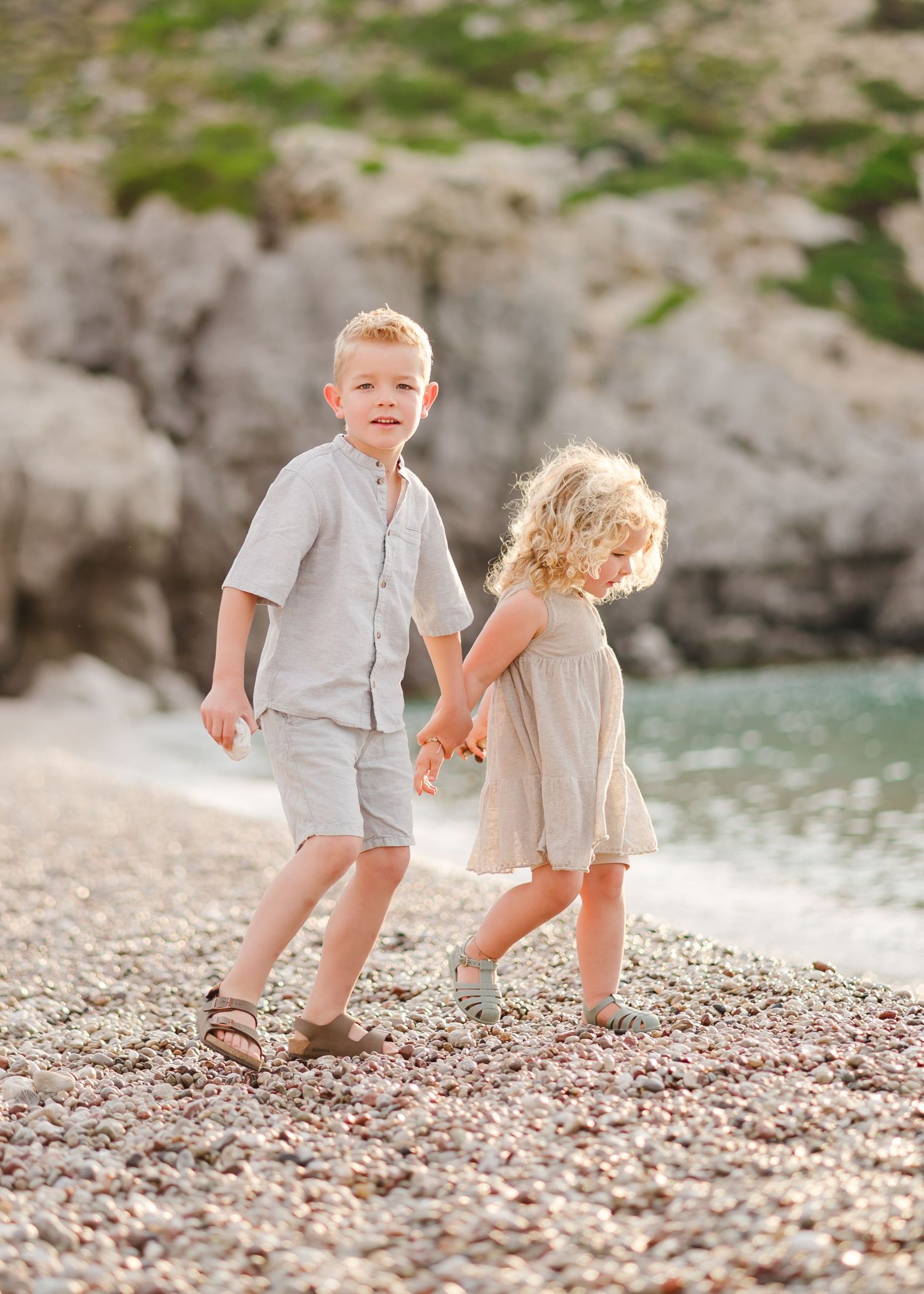 Two children hold hands while walking on a pebbly beach with cliffs and water in background. Fotograf Rhodos