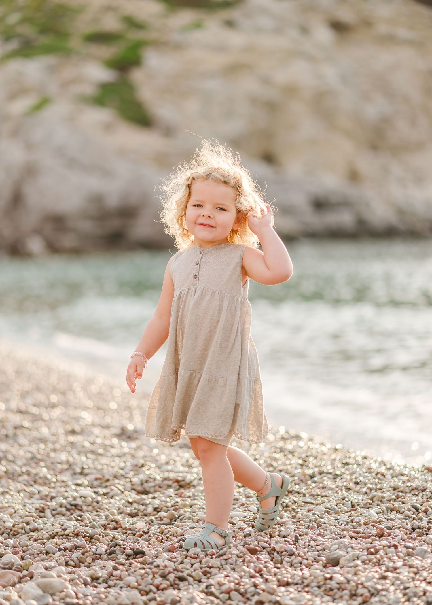 Smiling child in gray dress walks along rocky shoreline with mountains in background. Fotograf Rhodos