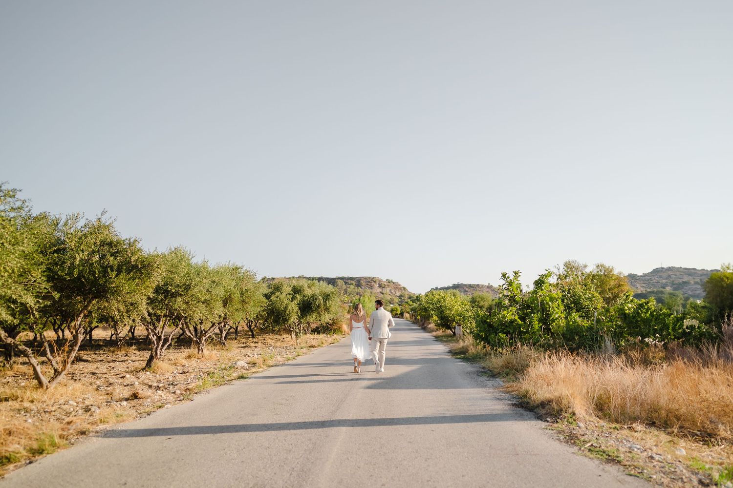 Paar in Hochzeitskleidung geht den ländlichen Weg entlang, umgeben von Weinberglandschaft. Fotografin Rhodos - Hochzeitsfotograf Rhodos Tina