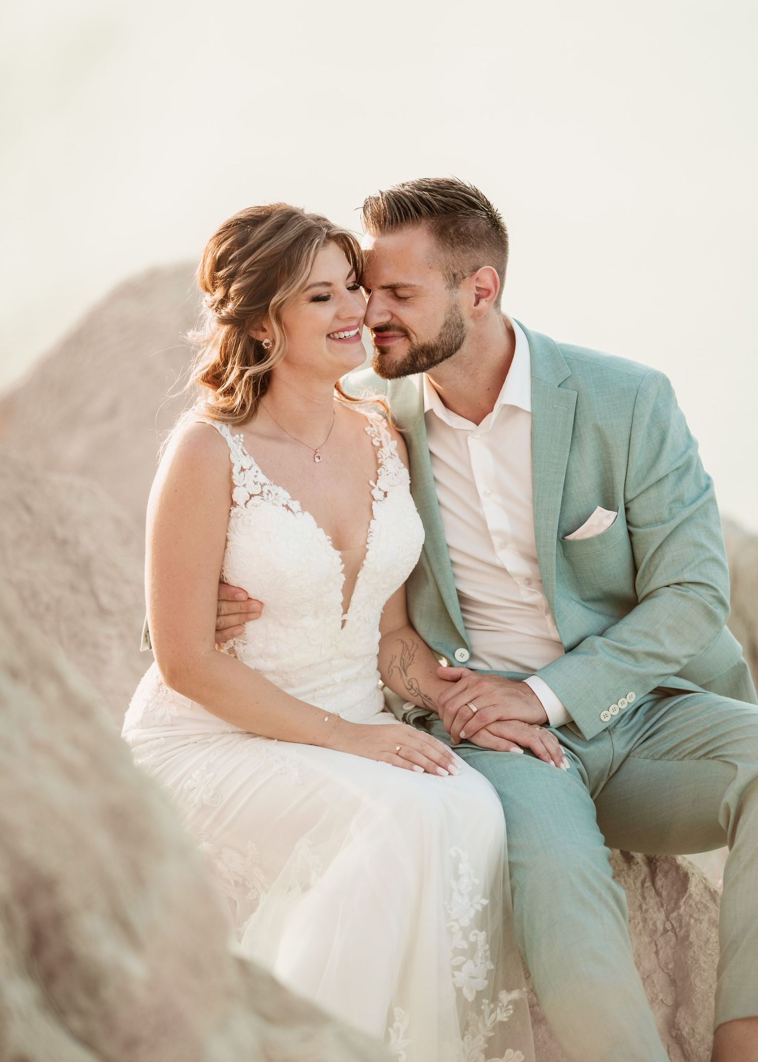 A romantic coastal wedding photo of a couple in mint green suit and white lace dress sitting tHochzeit Rhodos - Hochzeitsfoogether on rocks.