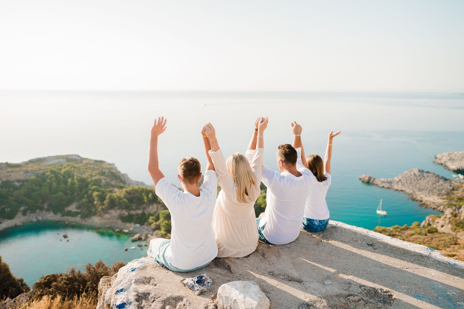 Group raising their arms in celebration while sitting on coastal cliff edge with scenic ocean view. Fotograf Rhodos