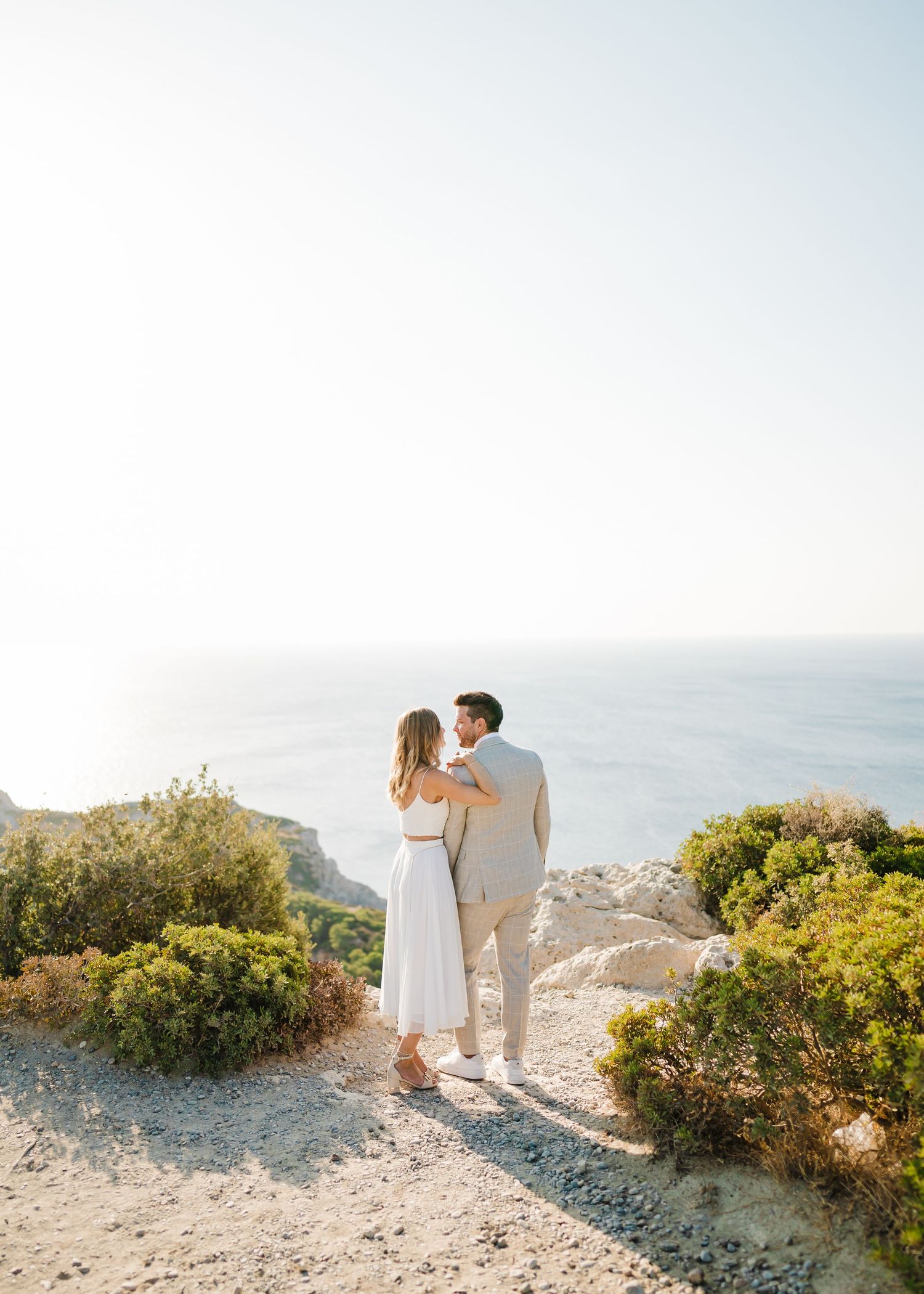 Ein Paar in leichter Sommerkleidung erlebt einen intimen Moment auf einem Klippenpfad an der Küste. Strandhochzeit Rhodos - Hochzeitsfotogra