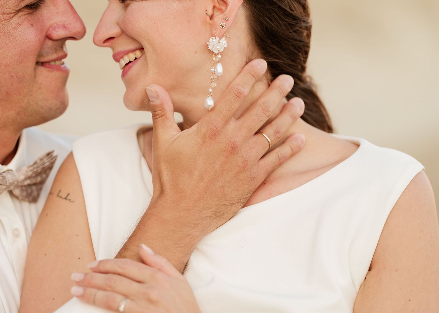 Nahaufnahme eines Paares in Weiß, das einen zarten romantischen Moment mit sanfter Beleuchtung und Perlentropfen-Ohrringen teilt. Hochzeiten