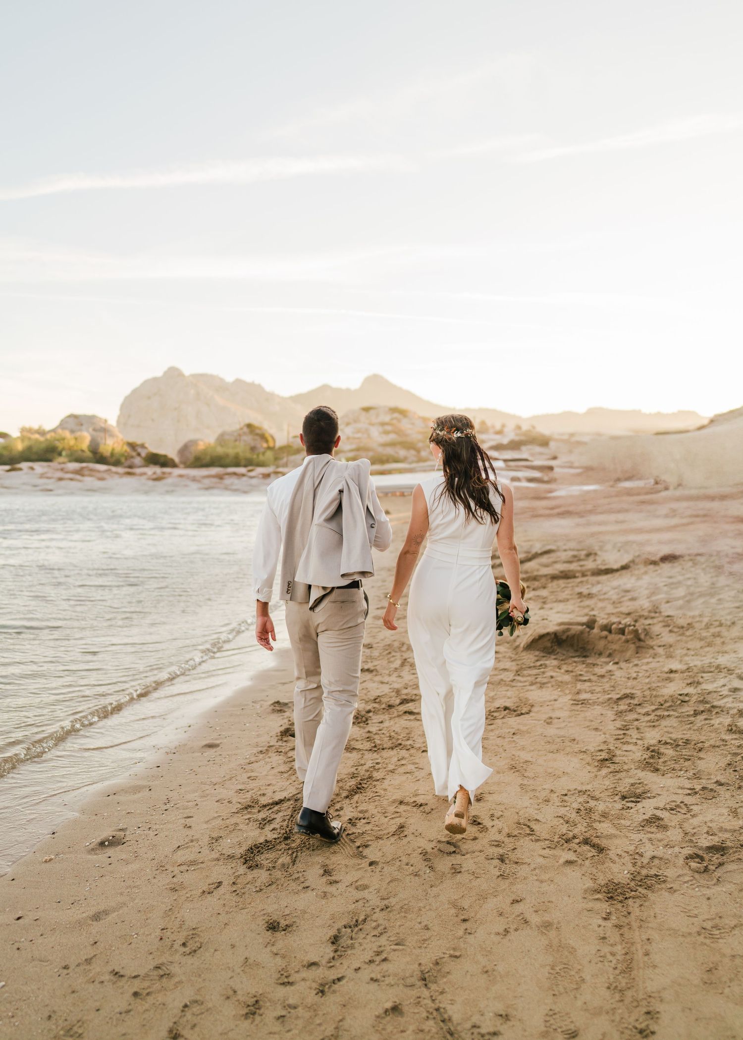 Zwei Menschen schlendern gemeinsam an einem Sandstrand mit fernen Bergen und Meerblick. Hochzeiten Griechenland - Hochzeitsfotograf Rhodos T