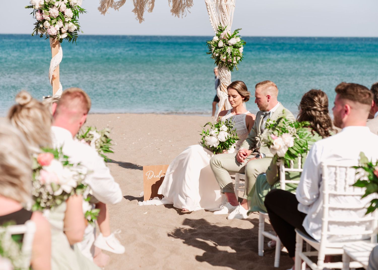 Wedding guests seated on white chairs during beach ceremony with floral decorations.