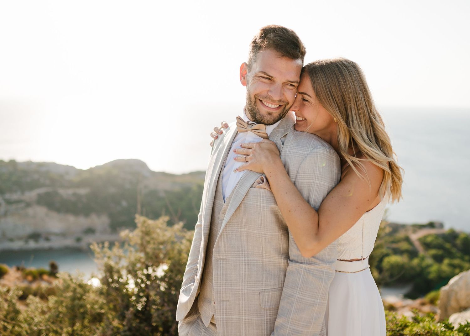Glückliches Paar in Hochzeitskleidung, das einen zarten Moment mit malerischem Blick auf die Küste teilt. Fotografin Rhodos - Hochzeitsfotog