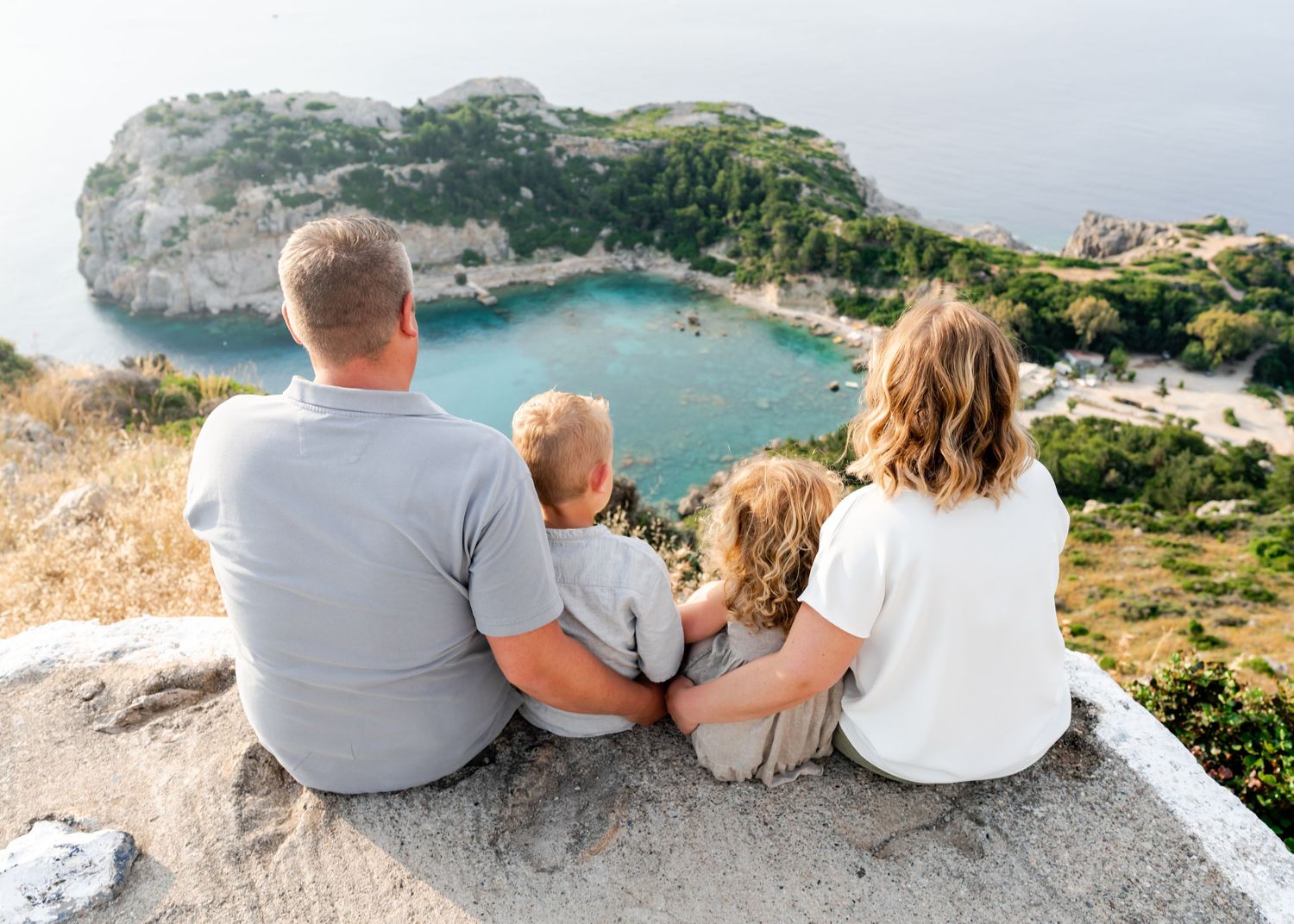Family sitting together on rocky coastal overlook enjoying scenic Mediterranean bay views. Fotograf Rhodos