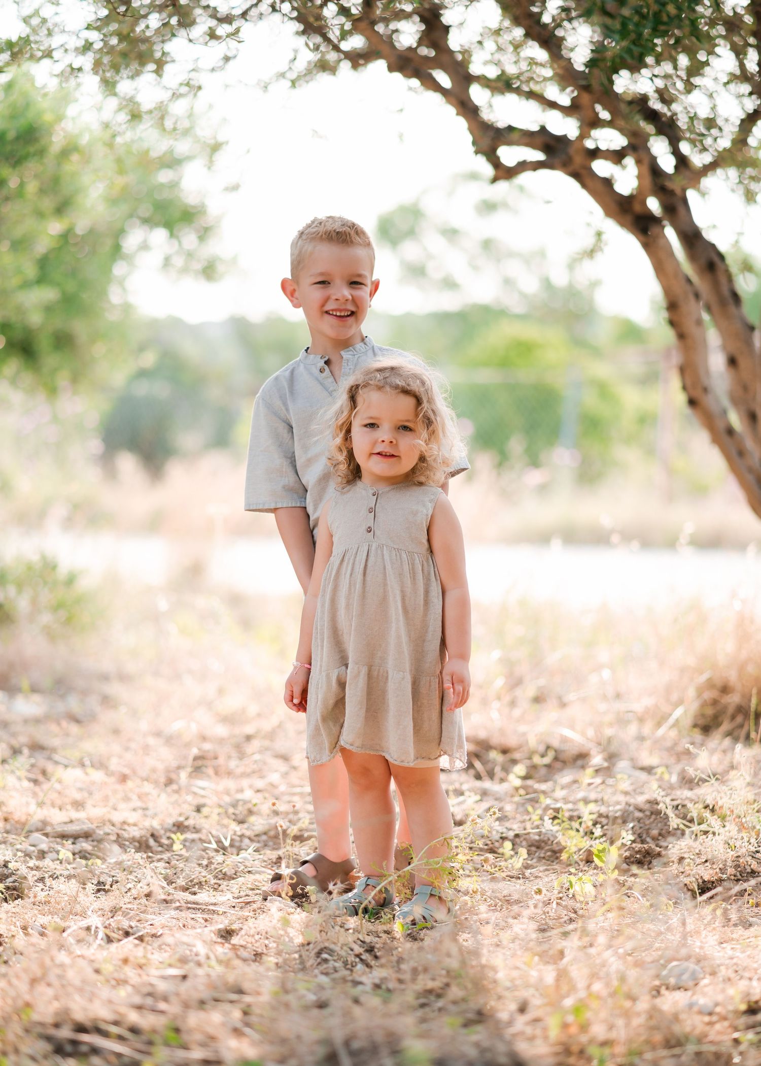 Two children stand together smiling in matching neutral-colored outfits under trees. Fotograf Rhodos