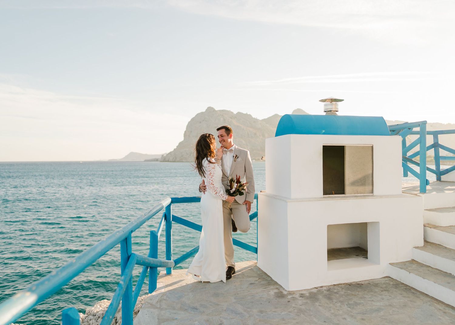 Hochzeitspaar, das sich auf weiße Küstenarchitektur mit blauen Geländern und Meerblick einlässt. heiraten am Strand - Hochzeitsfotograf Rhod