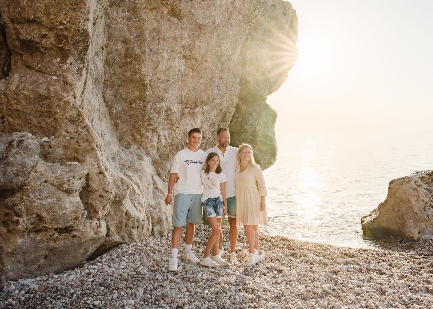 A group in white clothing poses together on a rocky beach against a bright sunset and stone wall backdrop. Fotograf Rhodos