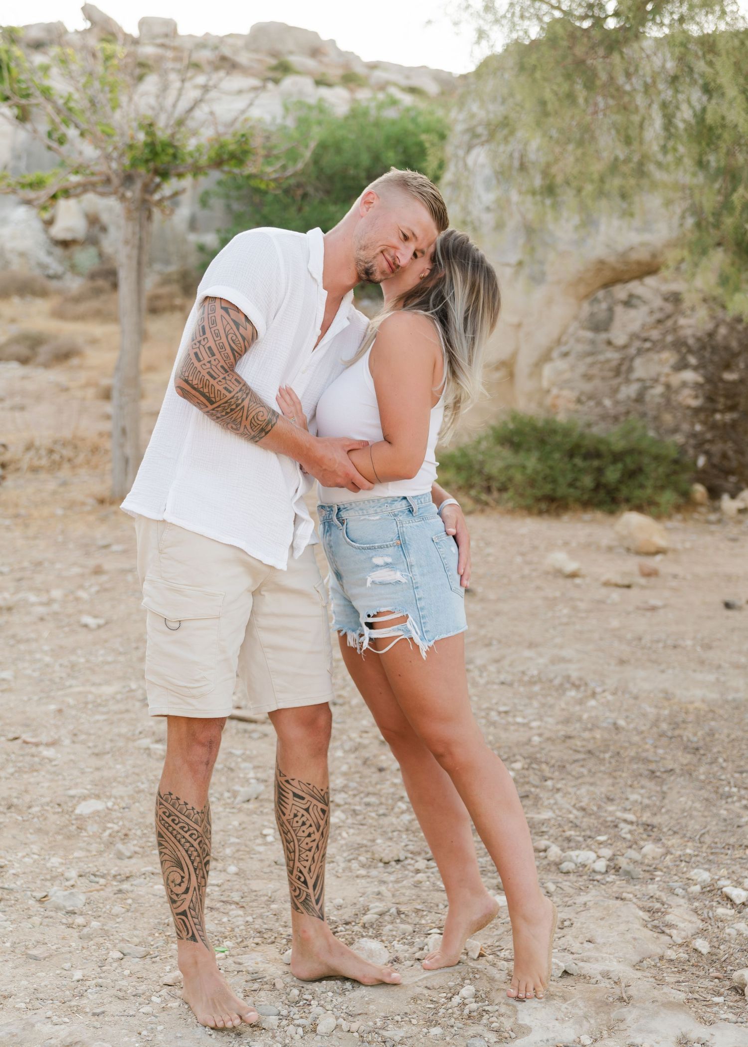 Barefoot couple embracing on desert terrain with visible tattoos and casual summer attire. Fotograf Rhodos