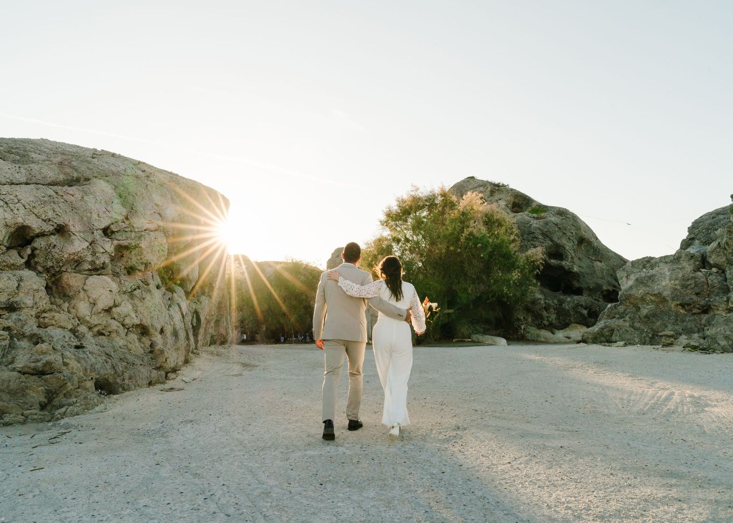 Paar tanzt während des Fotoshootings bei Sonnenuntergang freudig in weißen Outfits gegen Wüstenfelsen. Strandhochzeit Rhodos - Hochzeitsfoto