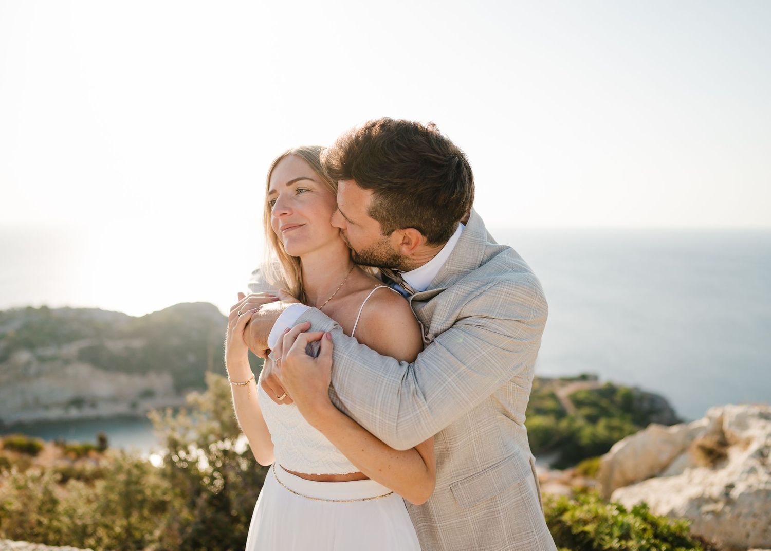 Paar in Hochzeitskleidung teilt intime Umarmung vor der Bergkulisse an der Küste. Fotografin Rhodos - Hochzeitsfotograf Rhodos Tina Lucius