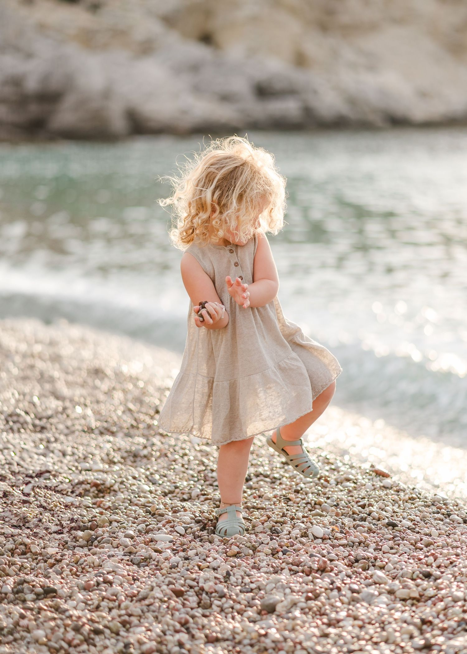 Young child in a beige dress twirls and plays on a rocky beach near the water. Fotograf Rhodos