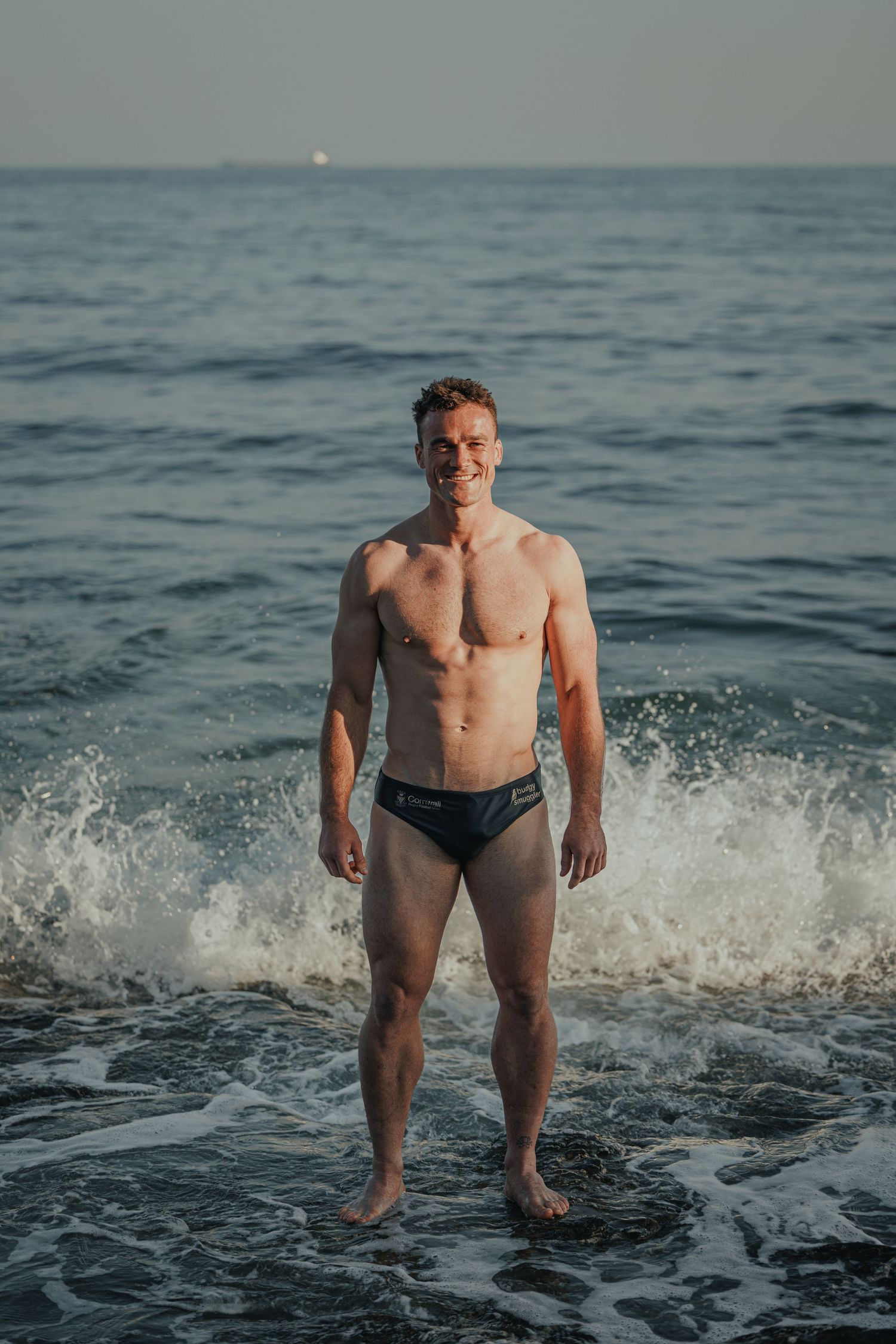 Athletic swimmer standing in crashing ocean waves at sunset.