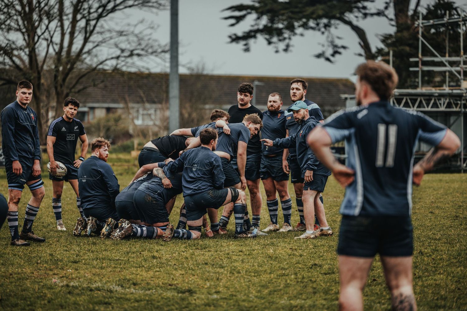 Rugby players gather in huddle formation while teammate observes from sideline.