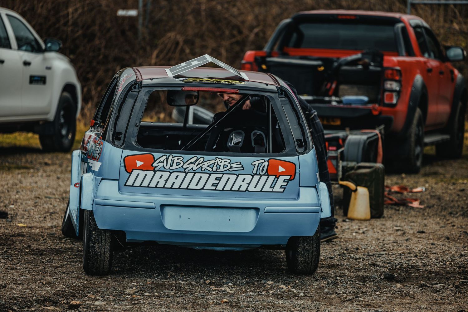 Light blue racing car with Mandeville branding parked on gravel lot.