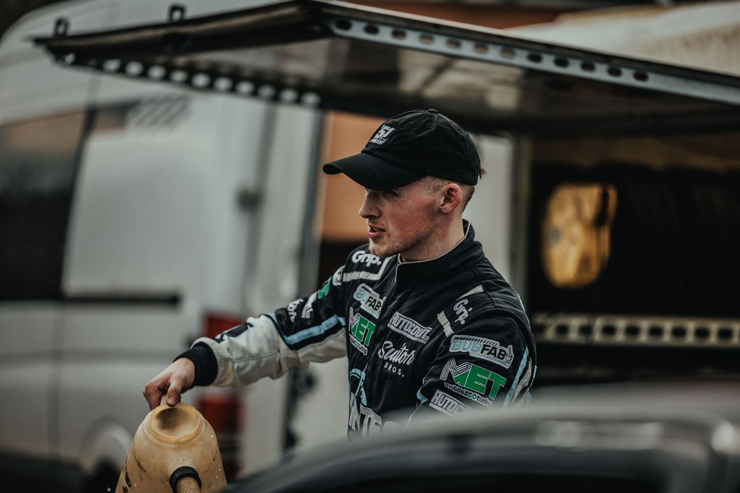 Racing driver in Vintec uniform reaching for equipment near transport vehicle.