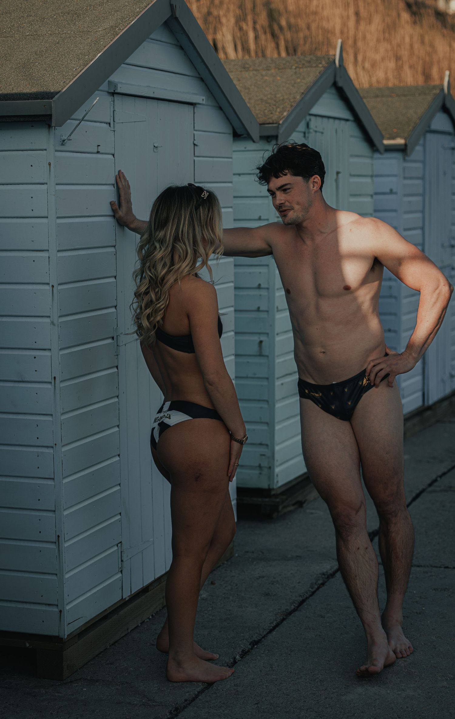 Athletic couple in swimwear posing playfully by beach huts.