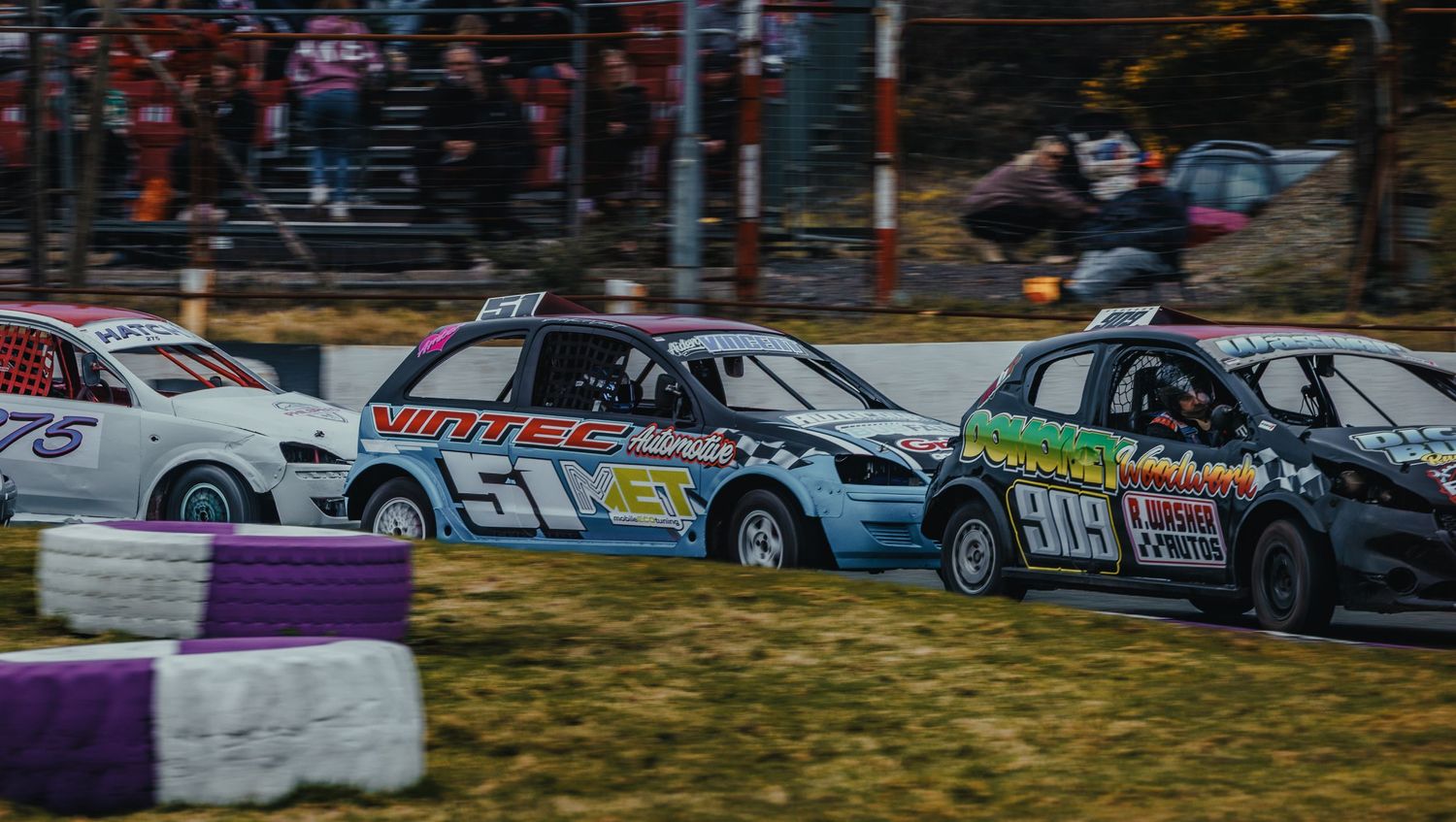 Race cars compete side by side on a dirt track with purple barriers during a stock car racing event.