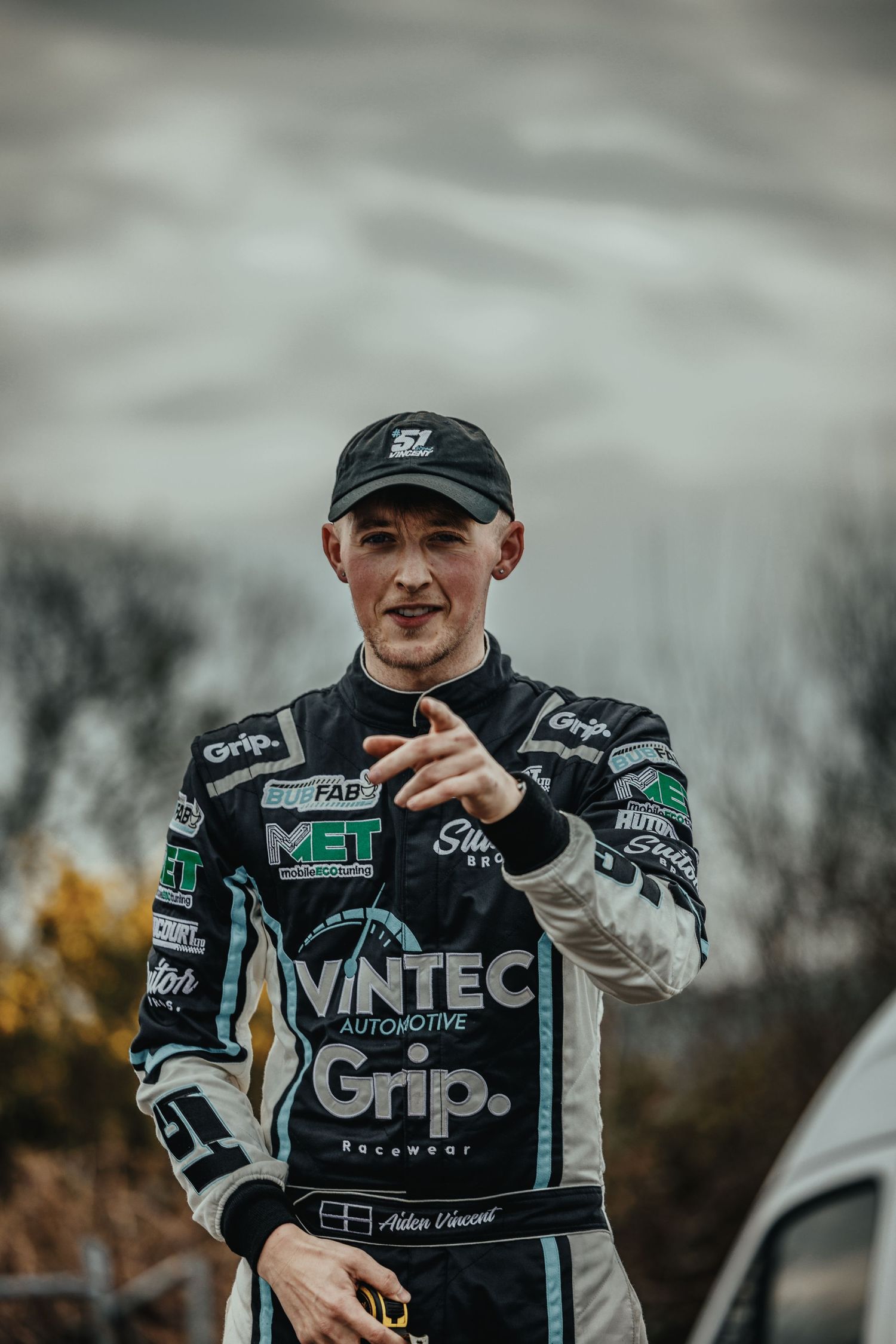 Racing driver in Vintec uniform gestures while standing outdoors against cloudy sky.