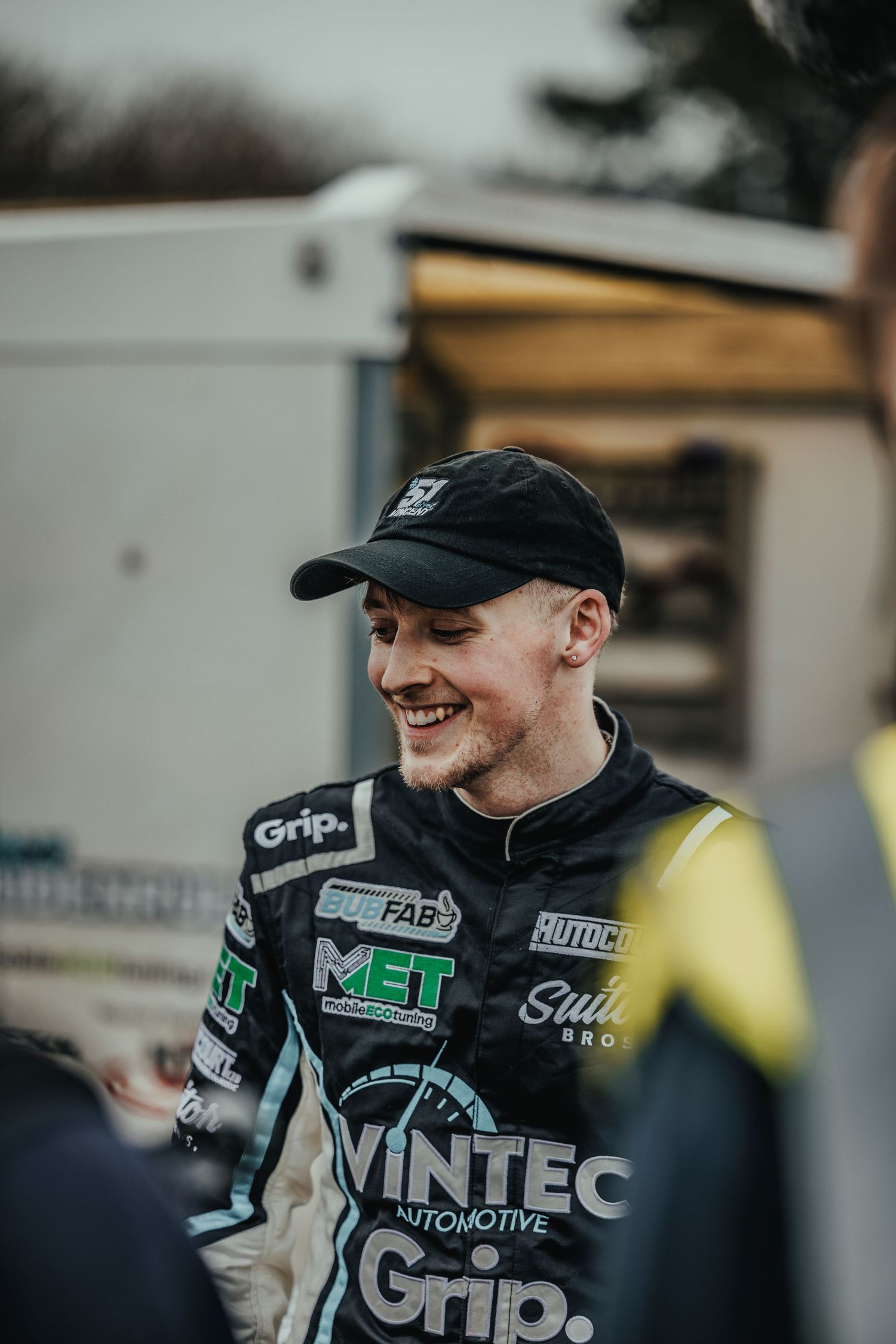 Race car driver in Vintec Automotive racing suit smiles while wearing a black baseball cap.
