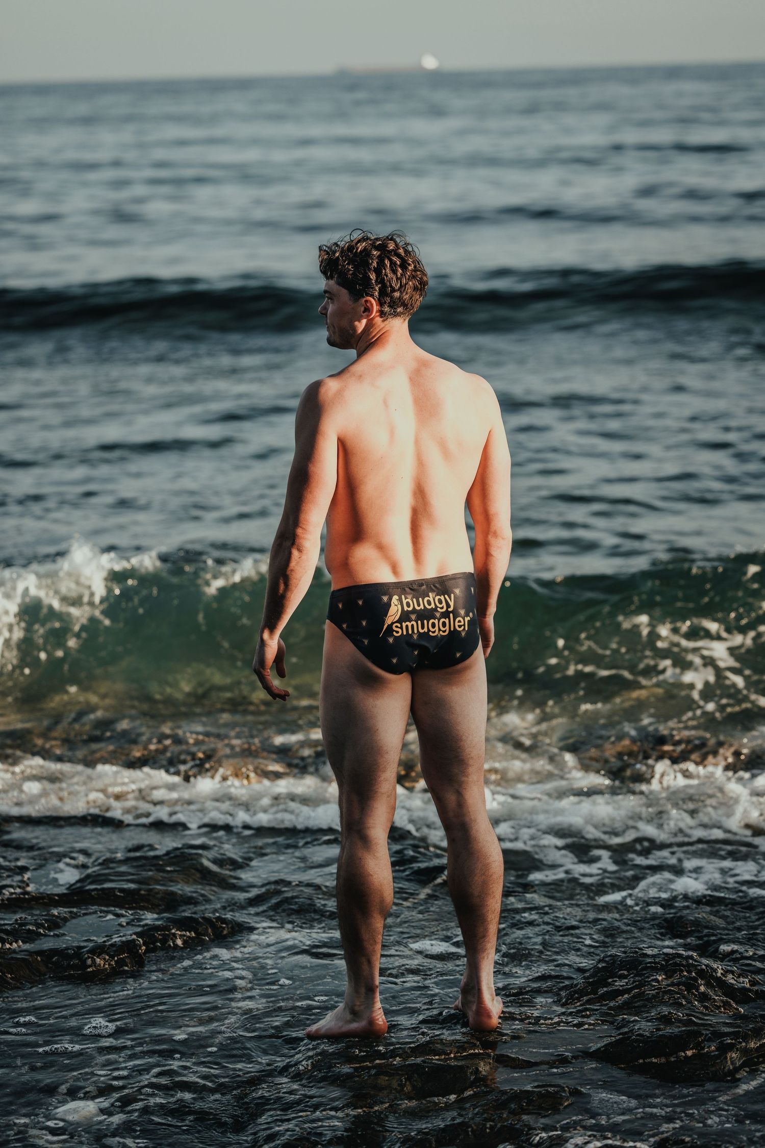 Person in swimwear stands facing the ocean waves at sunset.