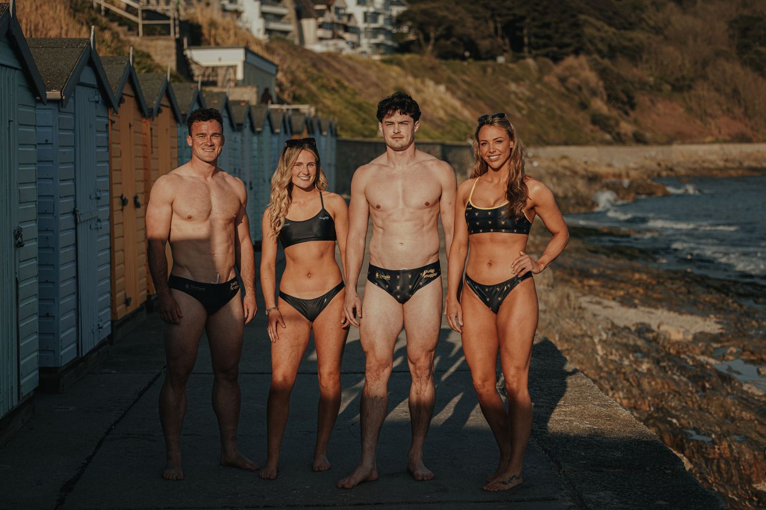 Group photo of friends in dark swimwear standing by coastal beach huts.