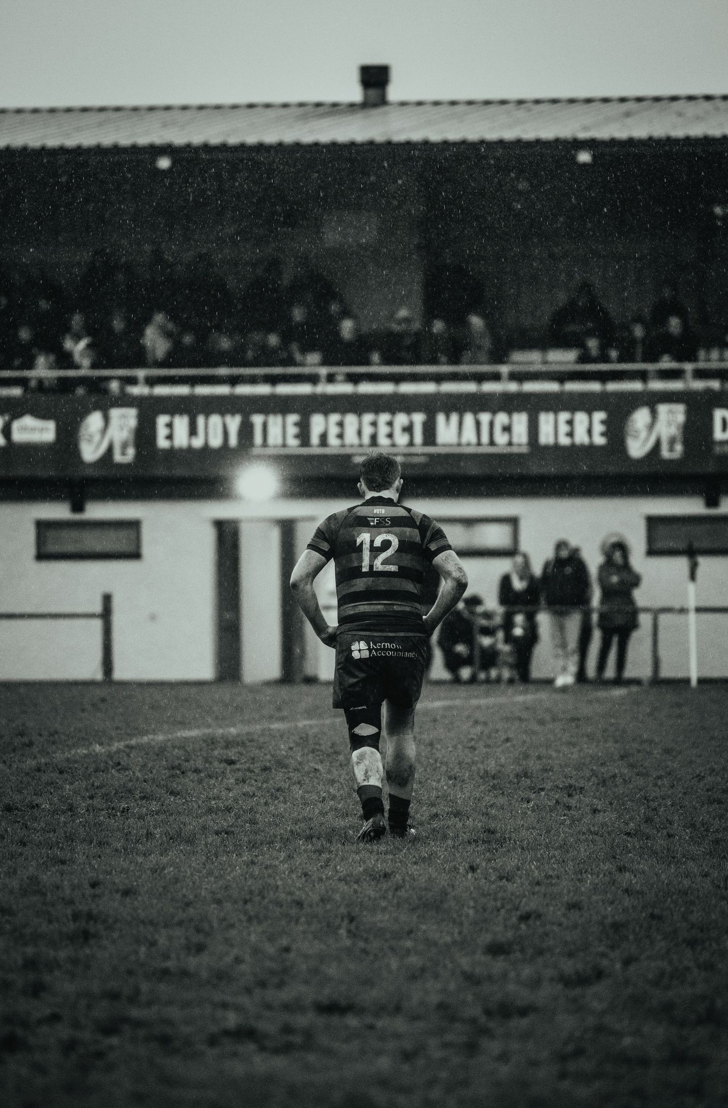 Rugby player wearing number 12 jersey walks across a rainy pitch with spectators and a stadium banner in the background.