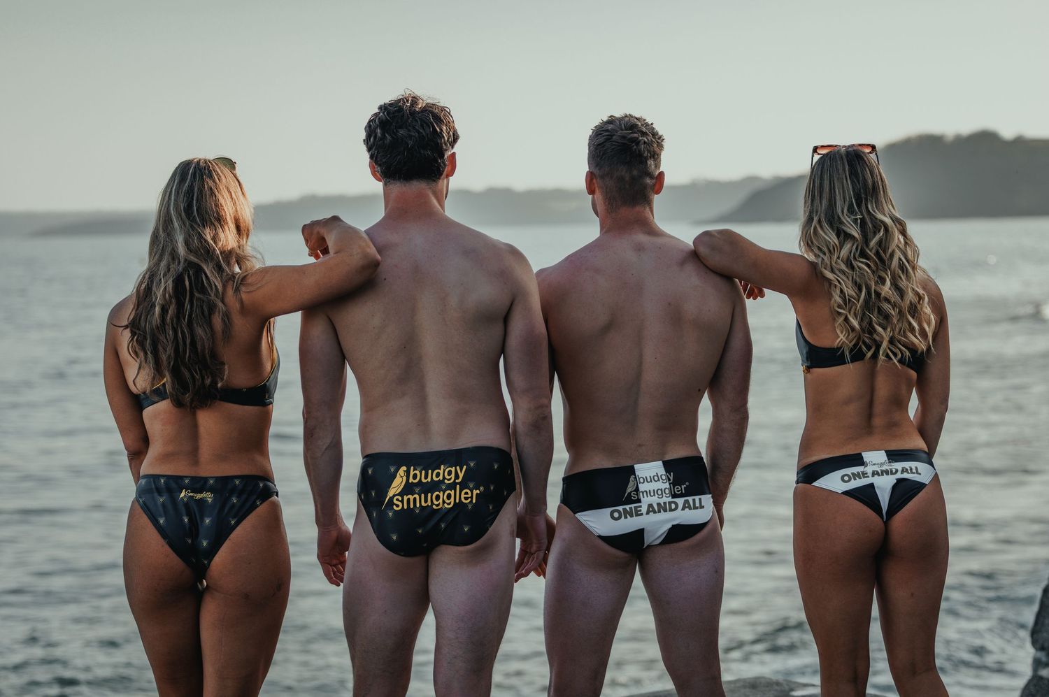 Four people in swimwear pose with humorous messages on their bottoms at the beach.