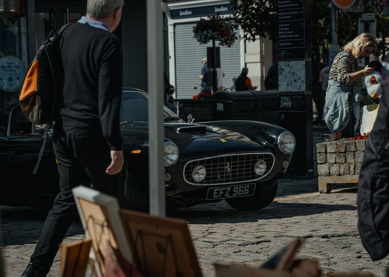 Classic vintage Ferrari parked on urban street with pedestrians walking by.