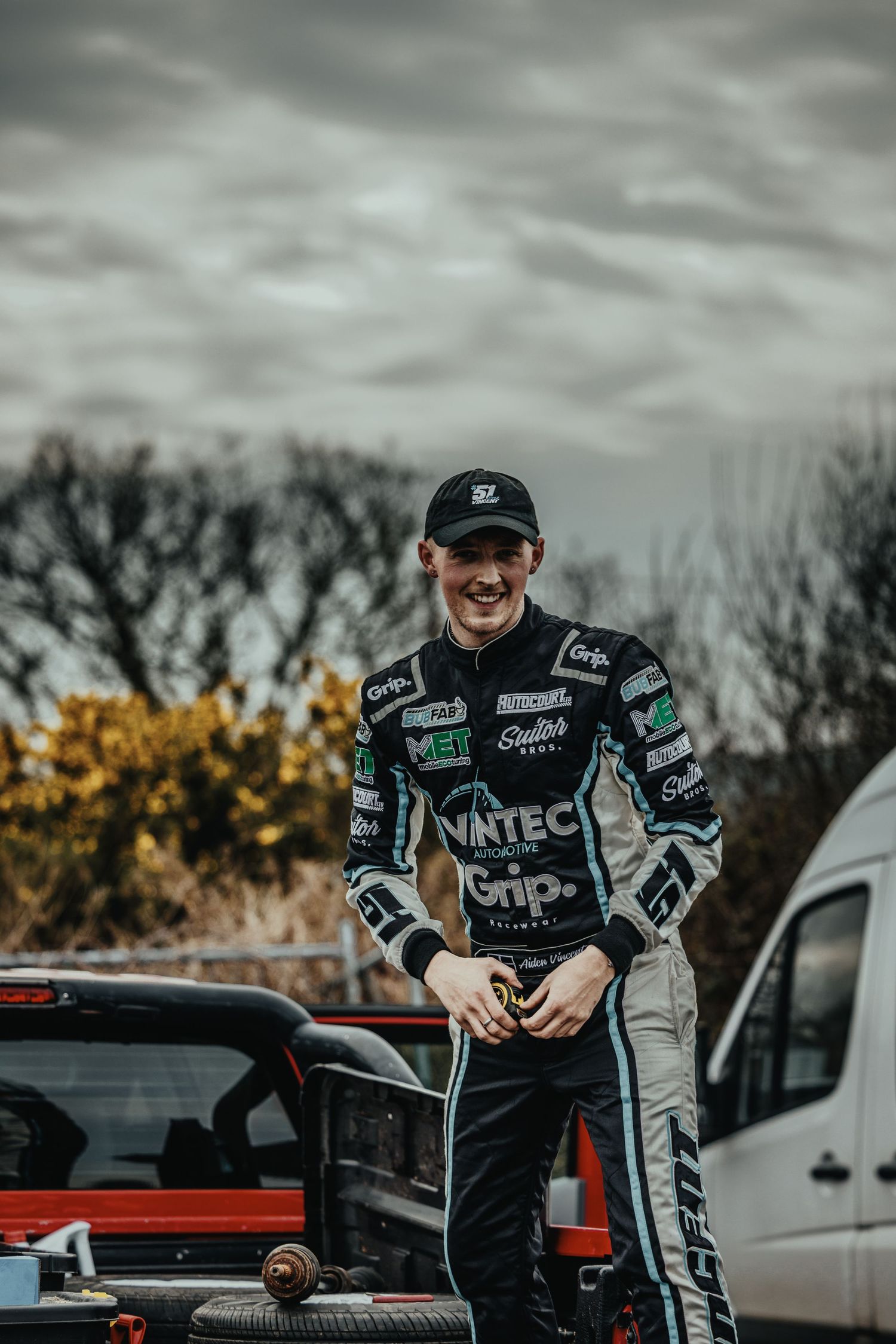 Motorsport athlete in Vintec gear smiles while standing between parked vehicles.
