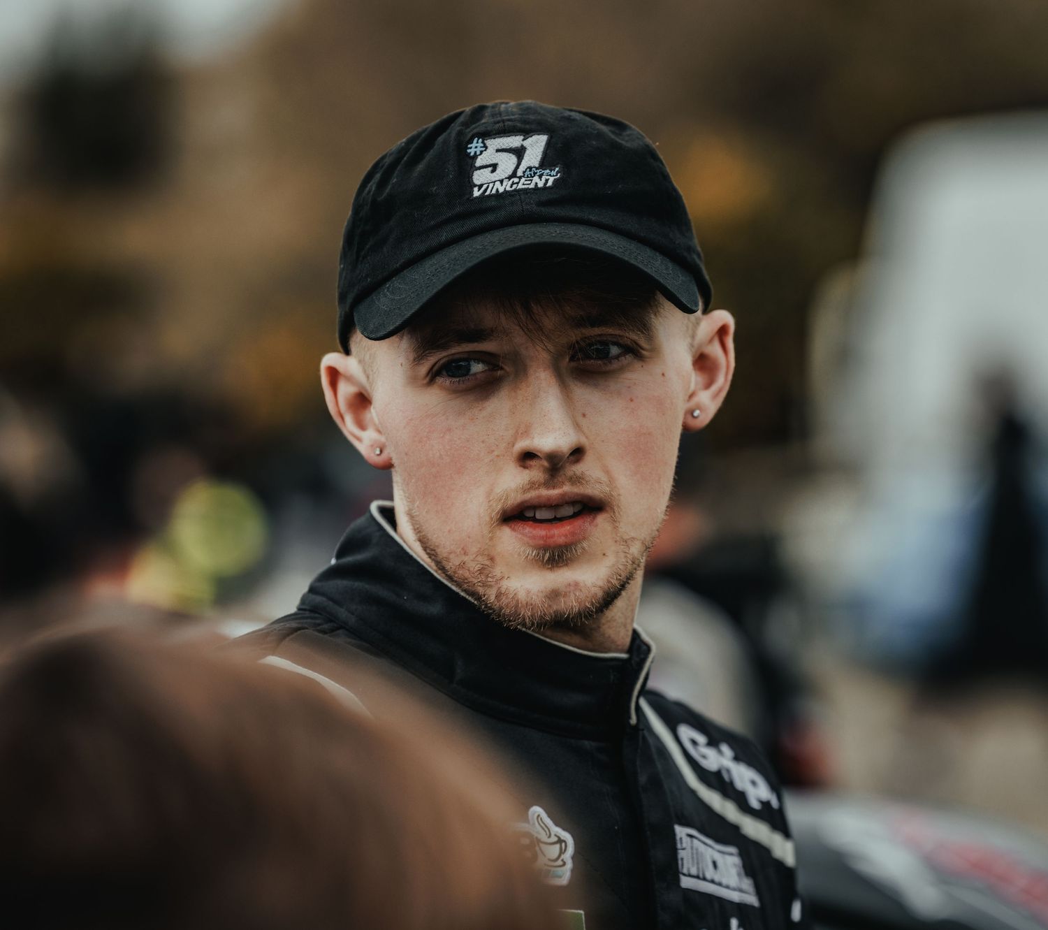 Racing driver in black cap and jacket at motorsport event.