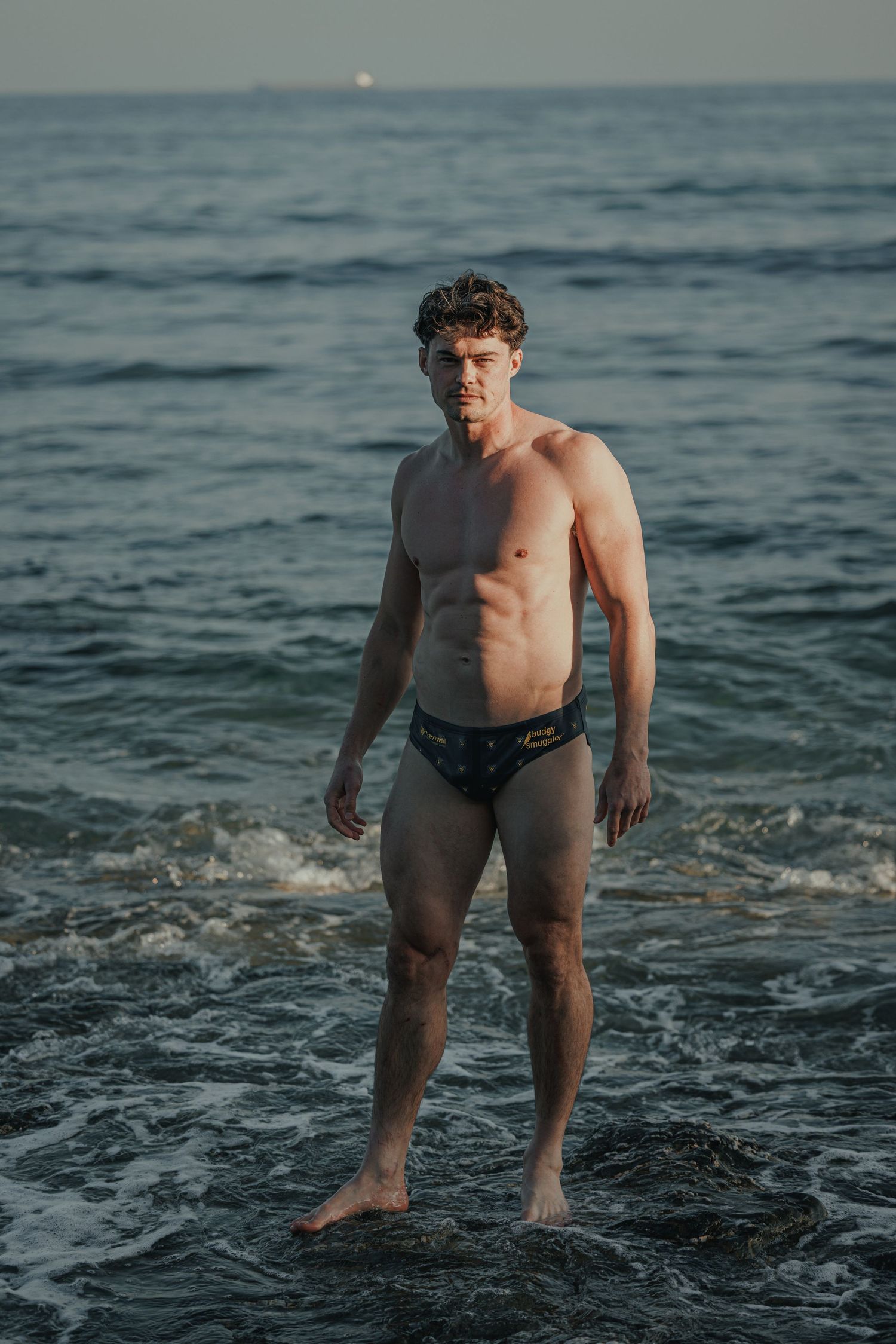 Athletic figure standing in ocean waves during sunset photoshoot.