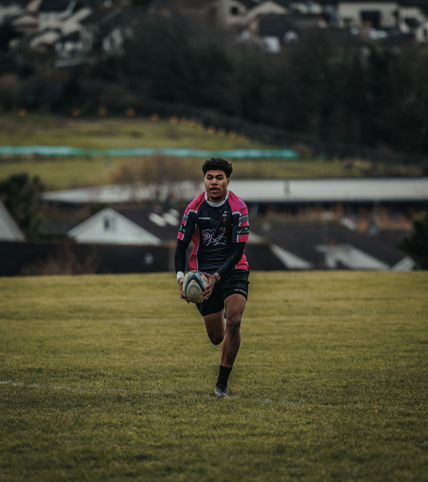 Rugby player running with ball across field with residential houses in background.