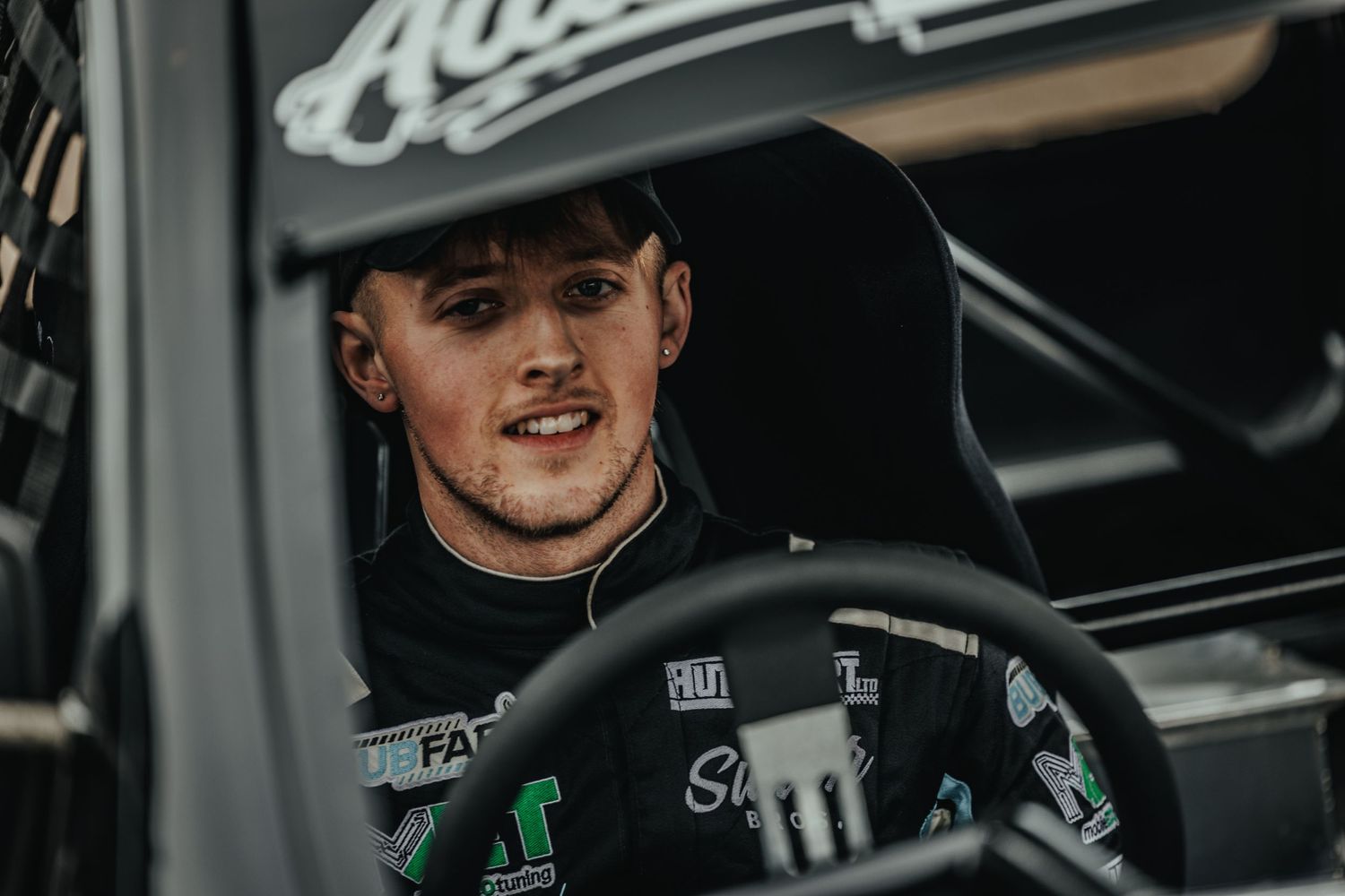 Professional driver sitting in racing car cockpit with sponsors and branding visible.