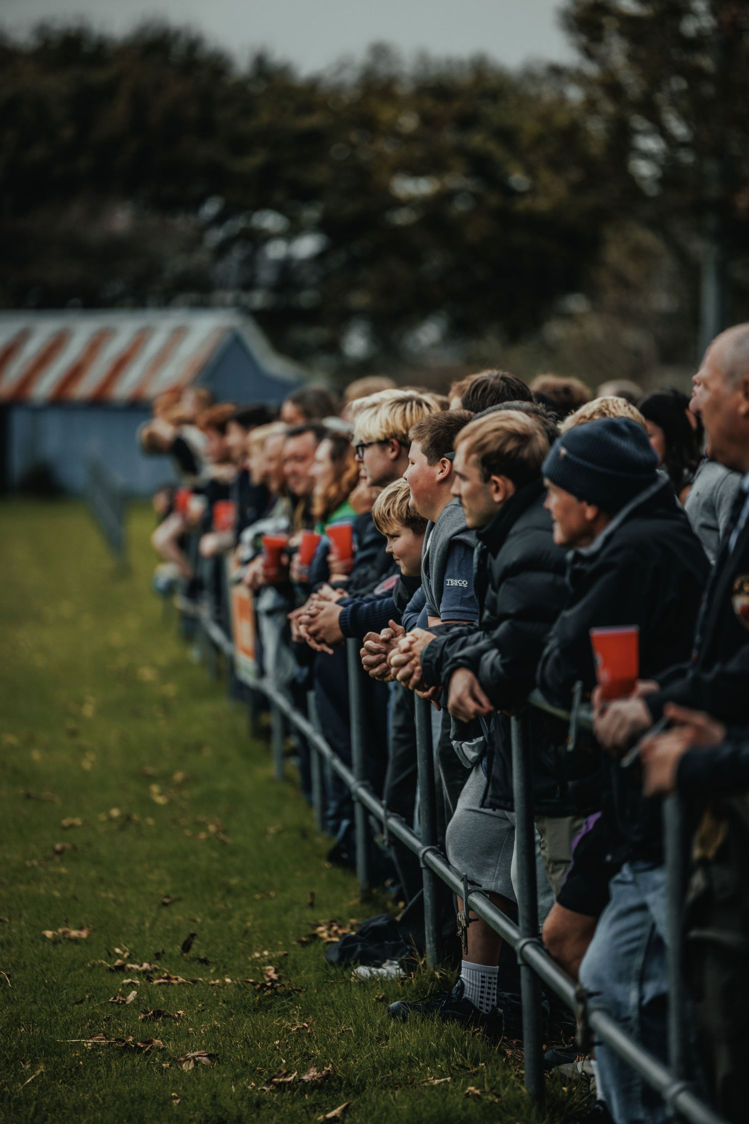 Crowd of spectators leaning on metal barriers watching a sports match outdoors on an overcast day.