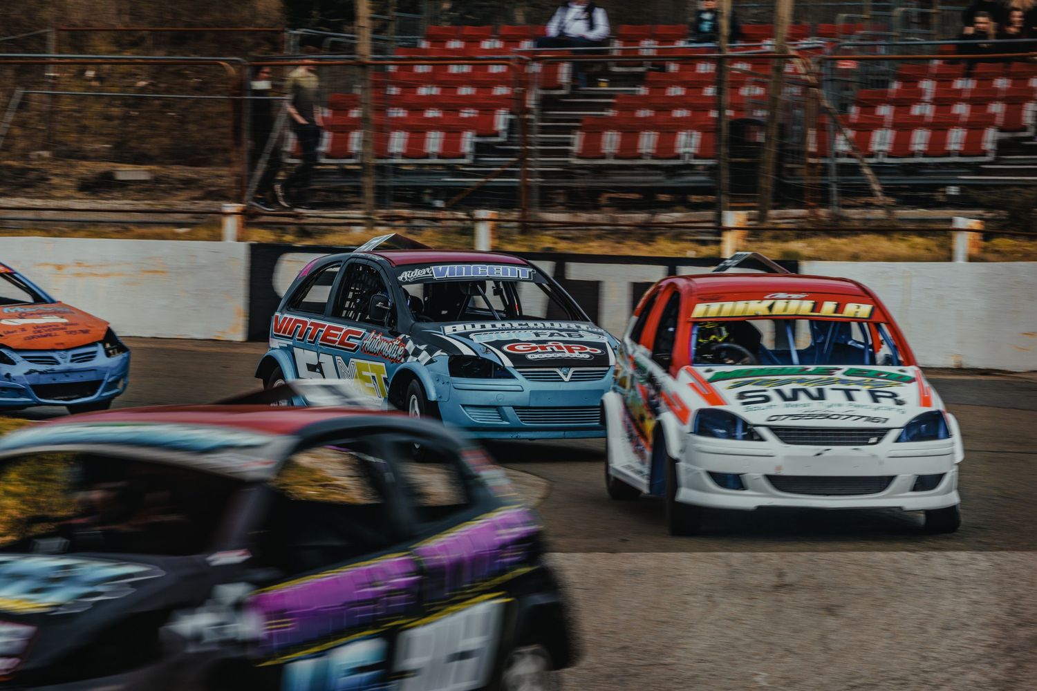 Multiple race cars competing closely together on an oval track with stadium seating visible.